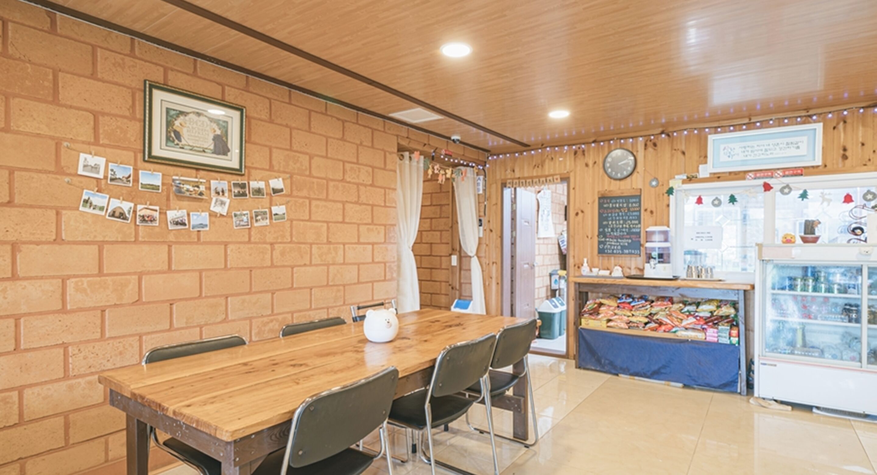 A restaurant with a brick wall, wooden ceiling, and a long wooden table with chairs. There is a refrigerator with various items on top and a chalkboard menu on the wall.