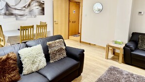 A modern living room with a black leather sofa, wooden chairs, a wooden bench, and a clock on the wall.