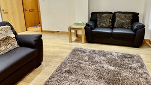 A living room with a black leather sofa, a wooden coffee table, a fluffy rug, a clock on the wall, and a door.