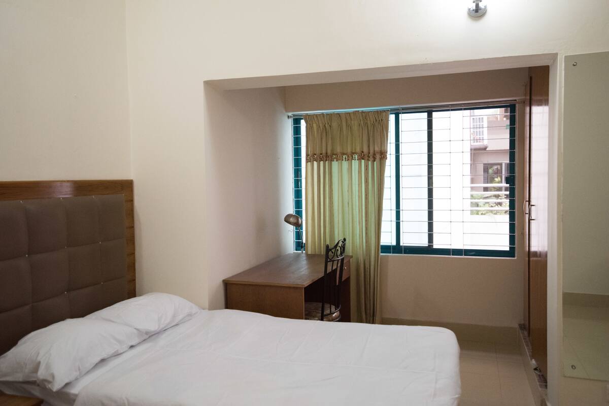 Une chambre à coucher avec un lit, un bureau et une chaise. Il y a une fenêtre avec des rideaux et une vue sur les bâtiments à l’extérieur.