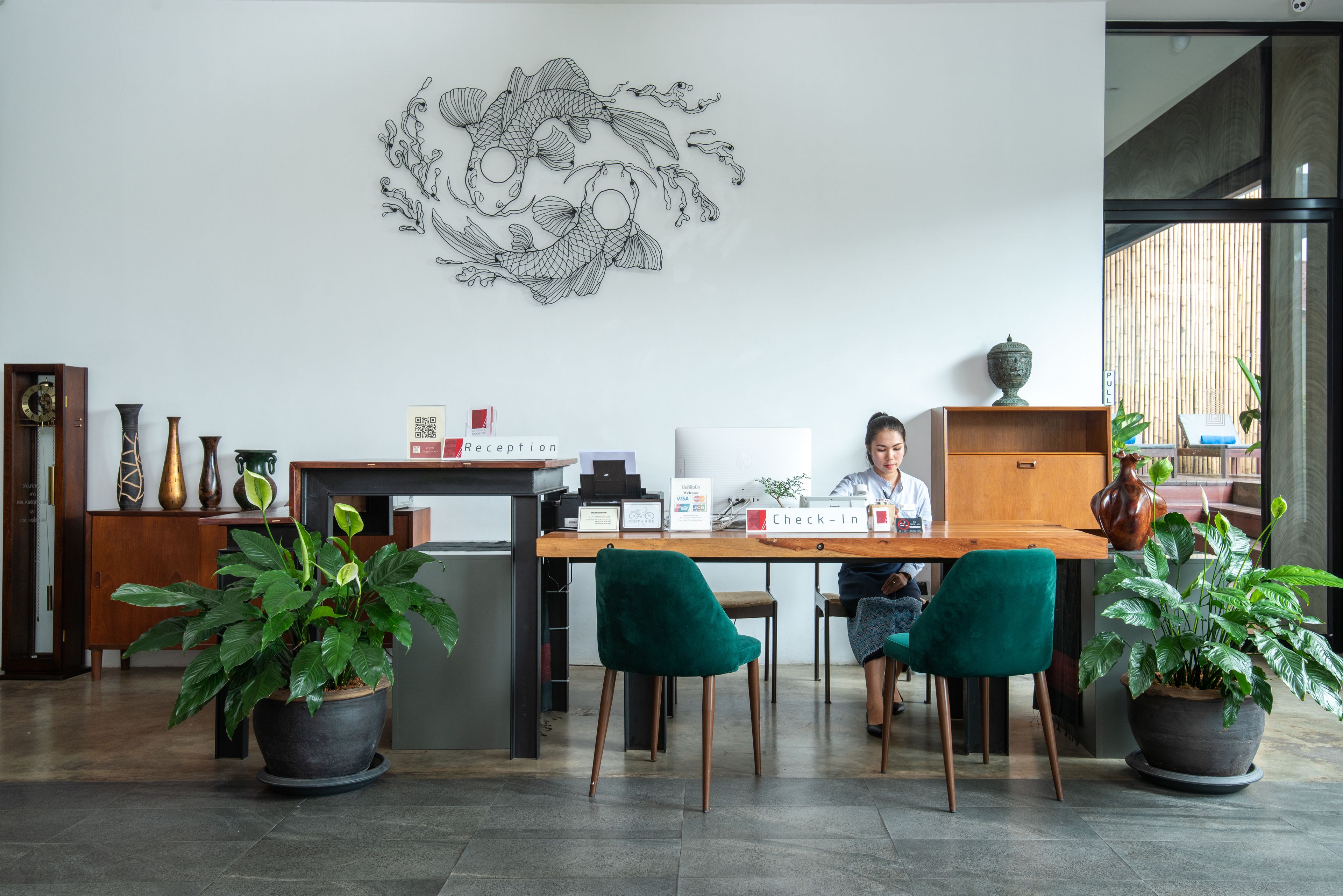 A reception desk with a nameplate, a check-in counter, and a receptionist. There are two potted plants, a decorative wall art, and a wooden cabinet with a vase.