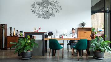 A reception desk with a nameplate, a check-in counter, and a receptionist. There are two potted plants, a decorative wall art, and a wooden cabinet with a vase.