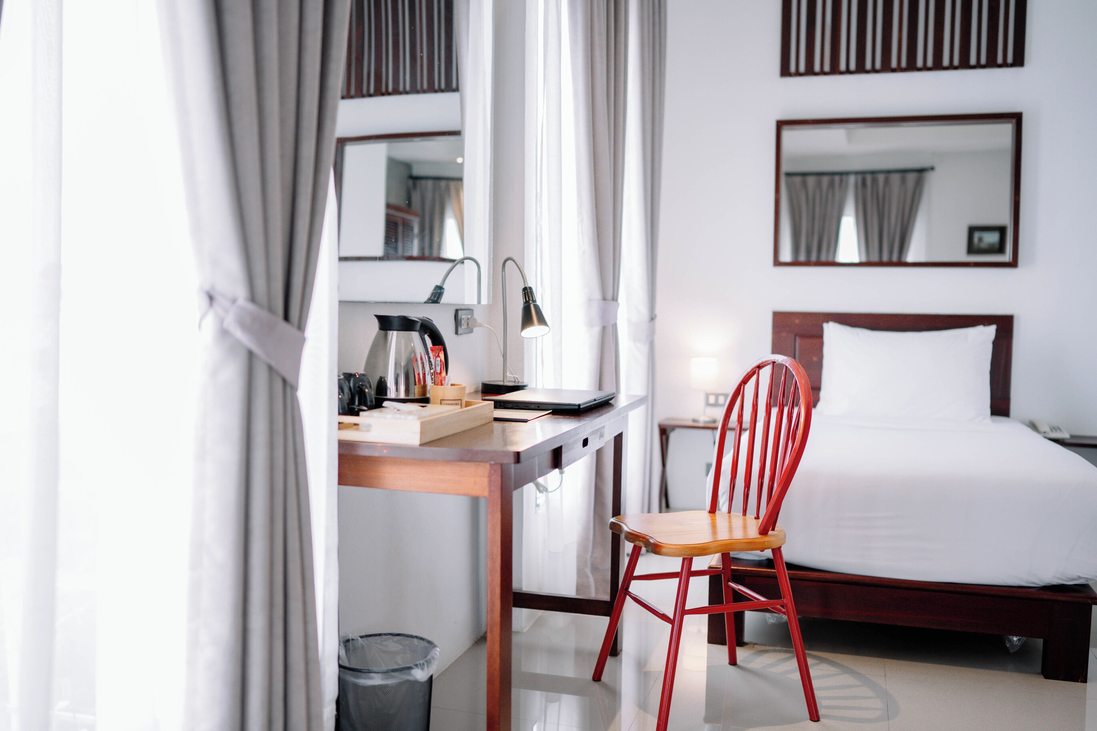 A hotel room with a bed, a desk with a laptop, a chair, a lamp, and a trash bin.
