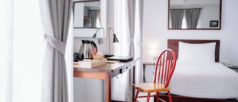 A hotel room with a bed, a desk with a laptop, a chair, a lamp, and a trash bin.