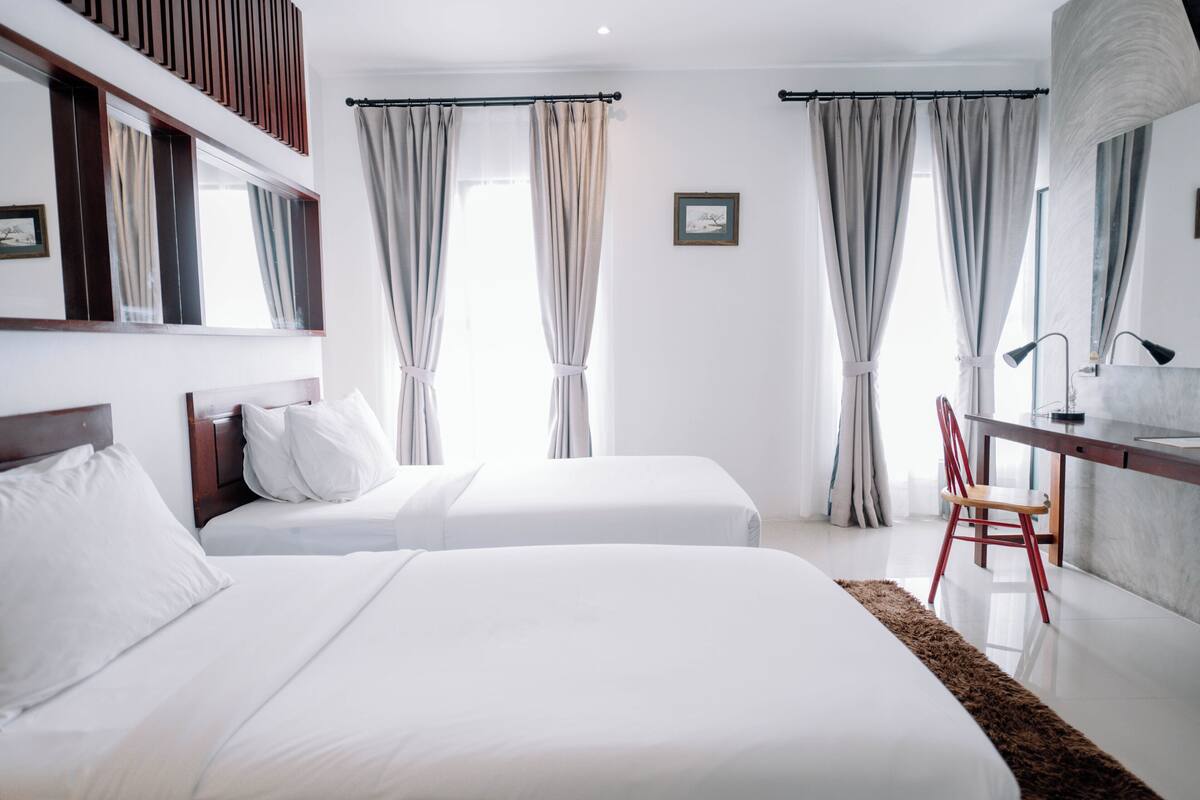 Habitación de hotel con dos camas, un escritorio con silla y un ventanal con cortinas.