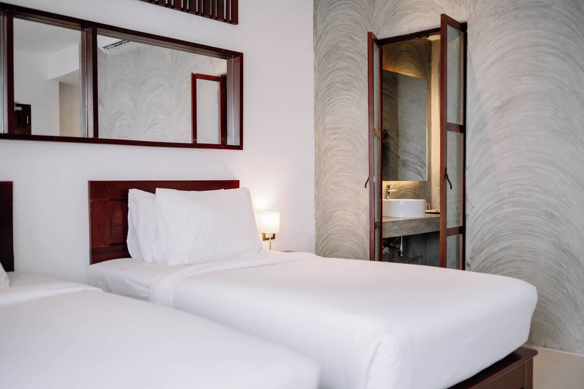 Habitación de hotel con dos camas, un espejo grande, un baño con lavamanos y una pared con textura.