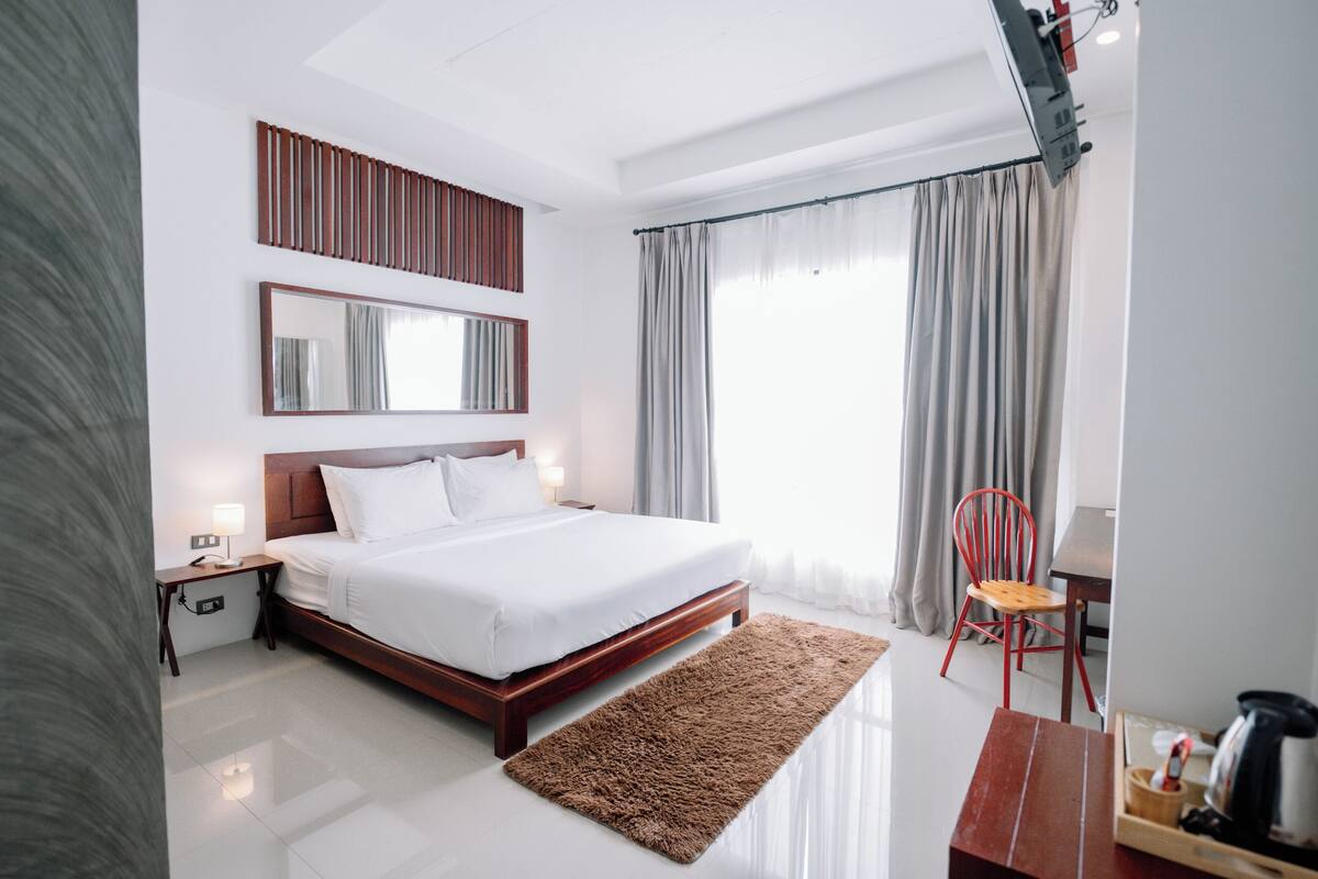 Habitación de hotel moderna con una cama grande, cabecera de madera, mesita de noche con lámpara, una silla roja y una pequeña mesa de madera con un hervidor.