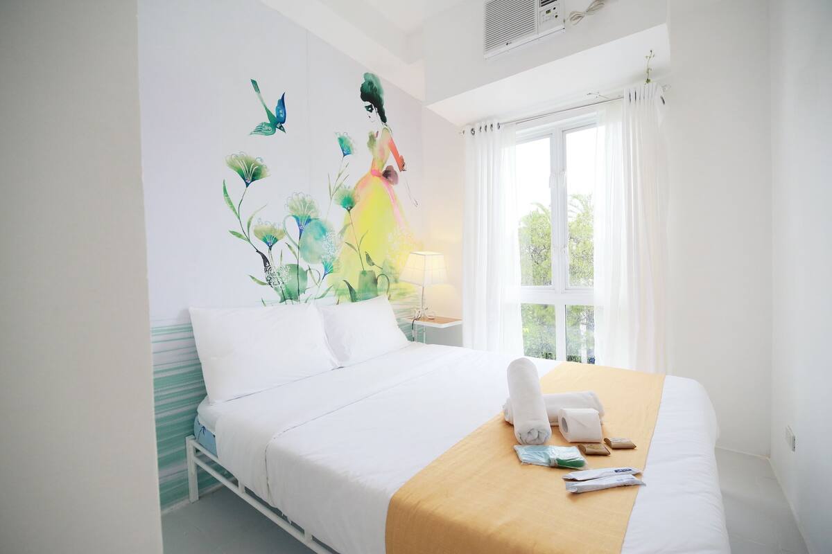 Habitación de hotel con una cama grande, una ventana con cortinas, un mural en la pared y una mesita de noche con lámpara.