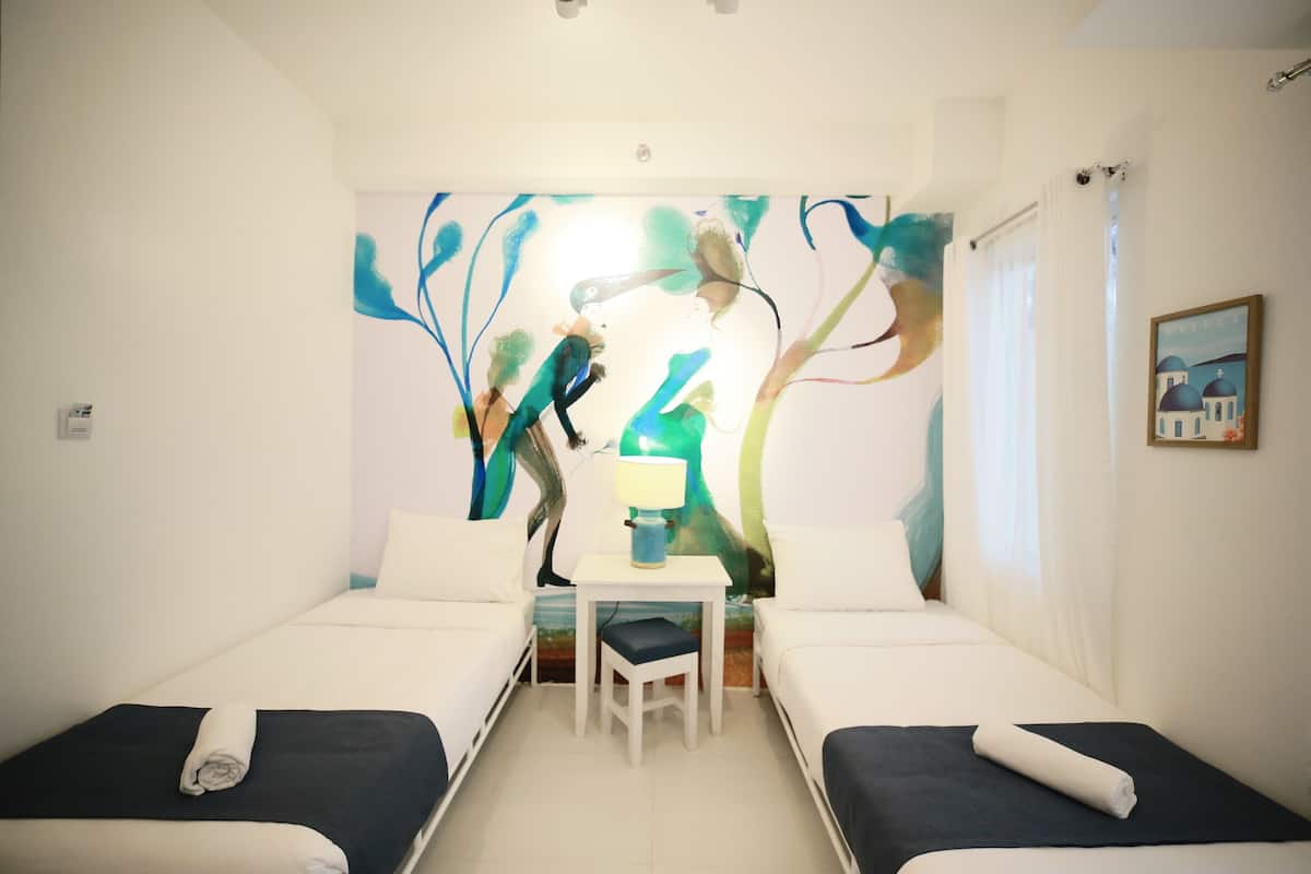 Een hotelkamer met twee enkele bedden, een kleine tafel met een stoel, en een kleurrijk abstract schilderij aan de muur.