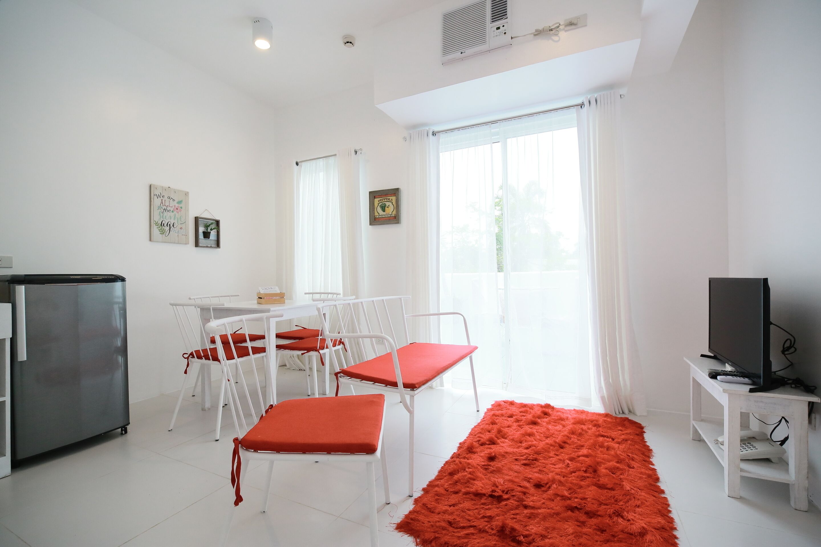 A bright room with white walls, a dining table with chairs, a red rug, a television on a stand, and a refrigerator.