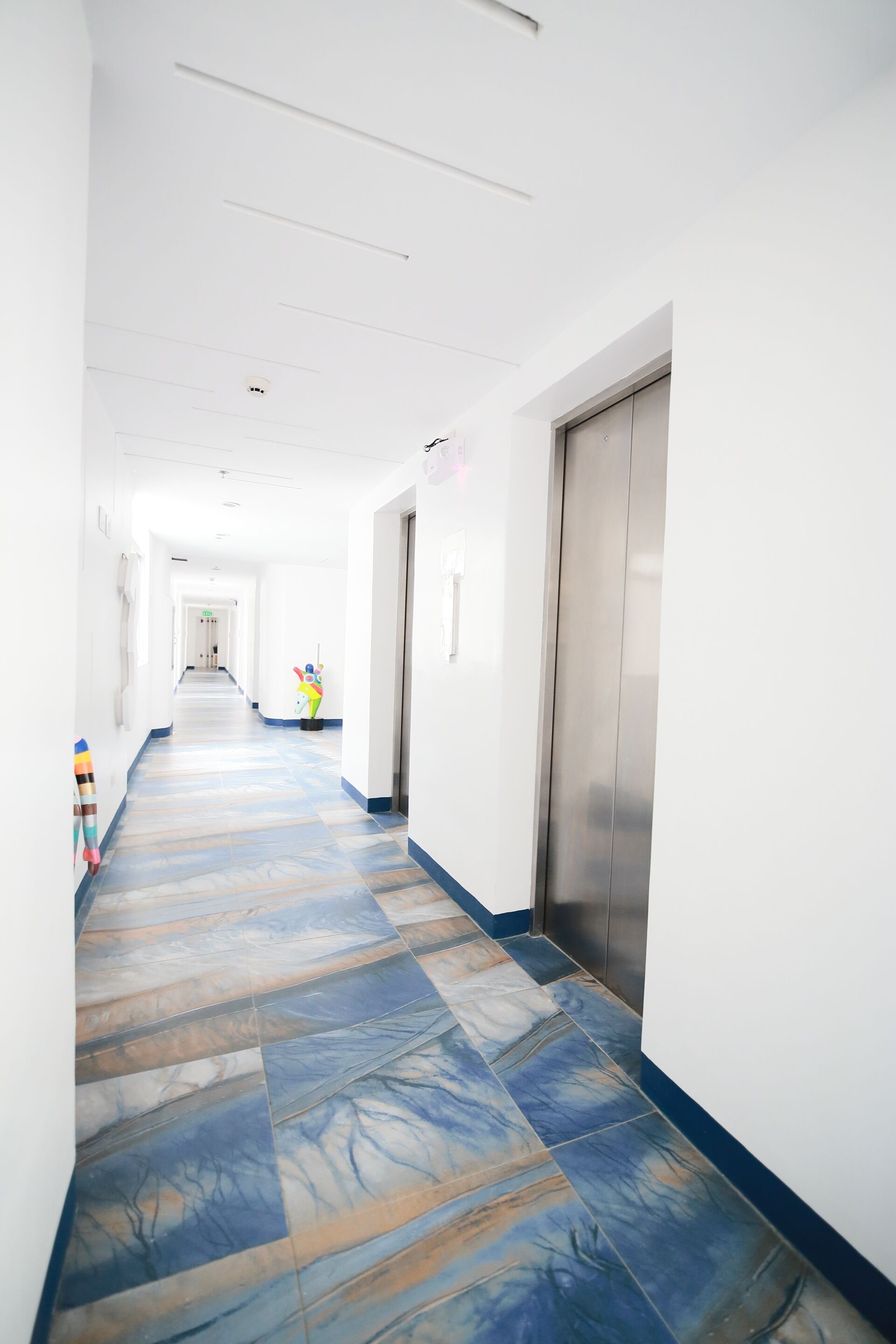 A long, narrow hallway with a patterned floor, white walls, and a metallic elevator on the right.
