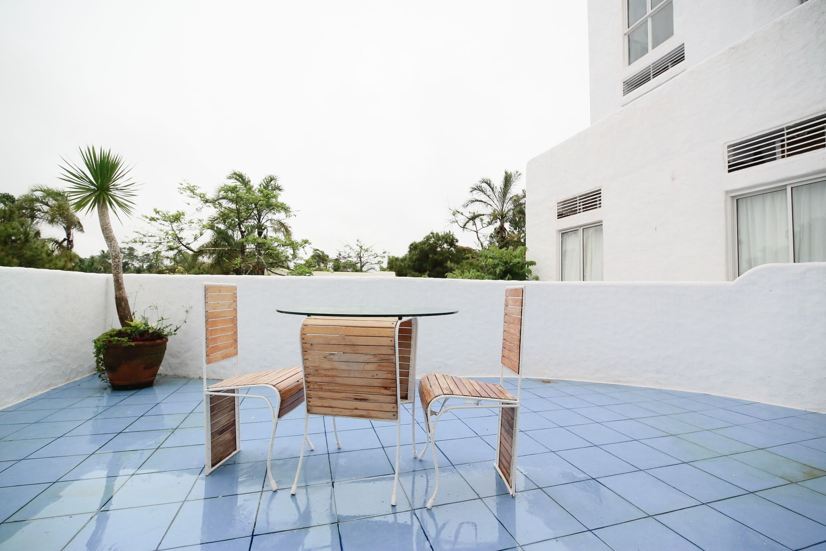 A patio with a round table and two wooden chairs, surrounded by a white wall and blue-tiled floor.