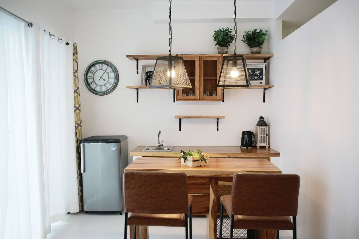 Una cocina moderna con una mesa de madera, dos sillas, lámparas colgantes, un reloj y estantes con plantas.