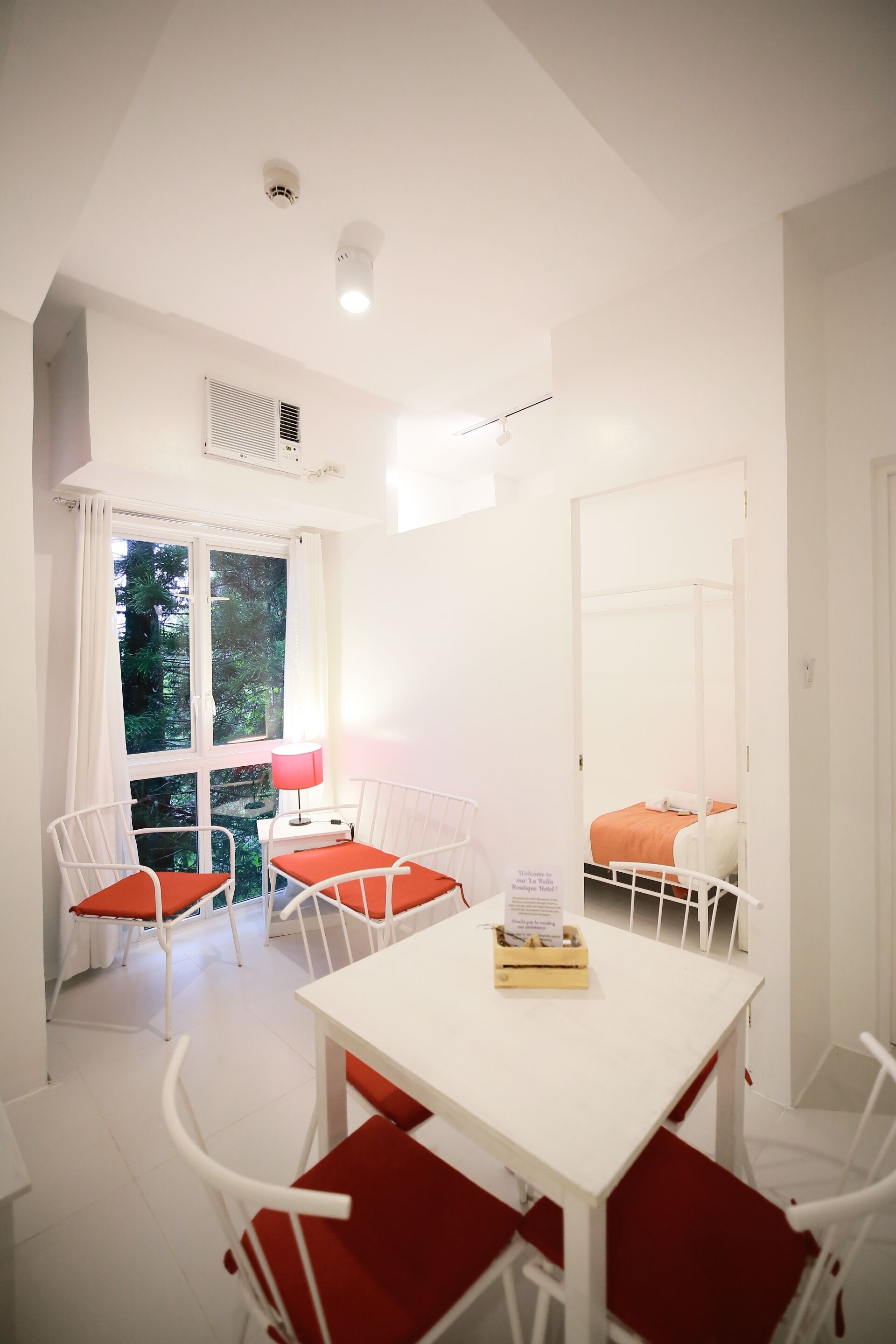 A bright room with white walls, a white table, and red chairs. There is a window with curtains, a bed with an orange blanket, and a white door.
