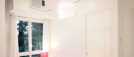 A bright room with white walls, a white table, and red chairs. There is a window with curtains, a bed with an orange blanket, and a white door.