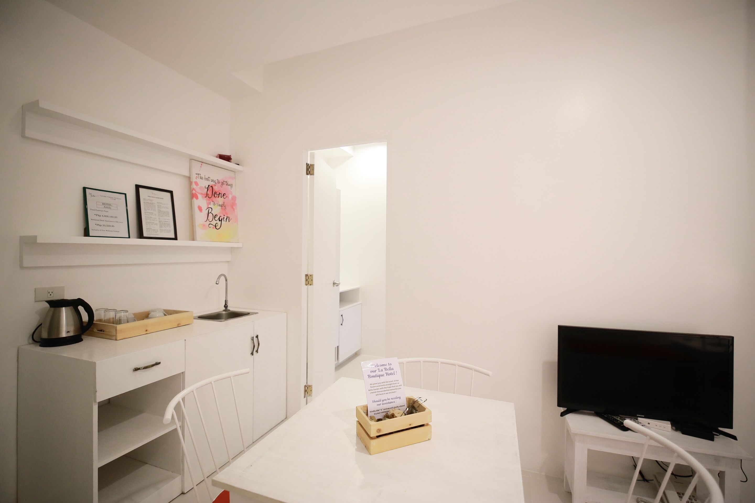 A small, white kitchenette with a coffee maker, a kettle, and a tray of glasses on the counter. There is a sign on the table and a television on a stand to the right.