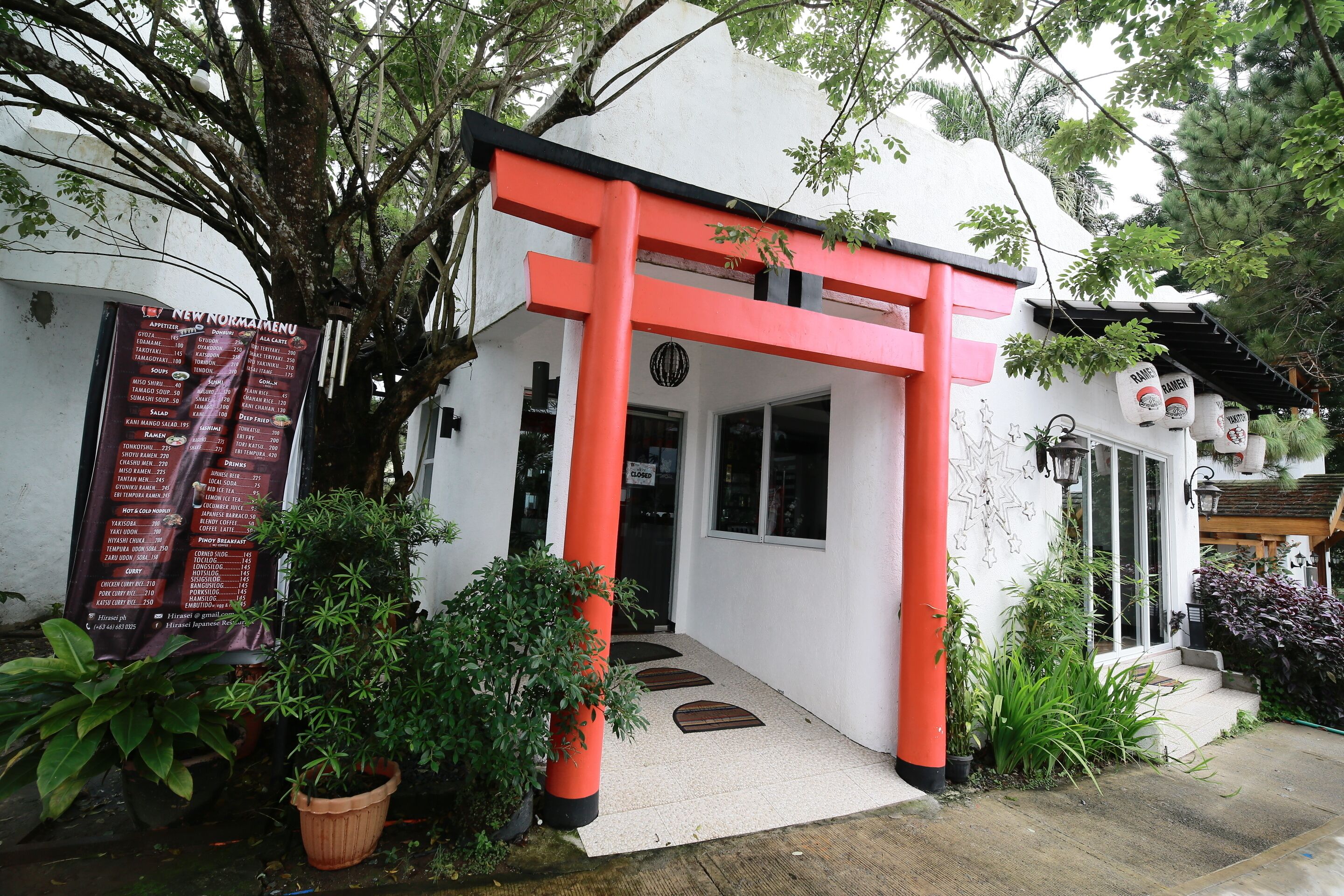 Un edificio con un arco de torii rojo, un cartel con el menú afuera y plantas en macetas alrededor.