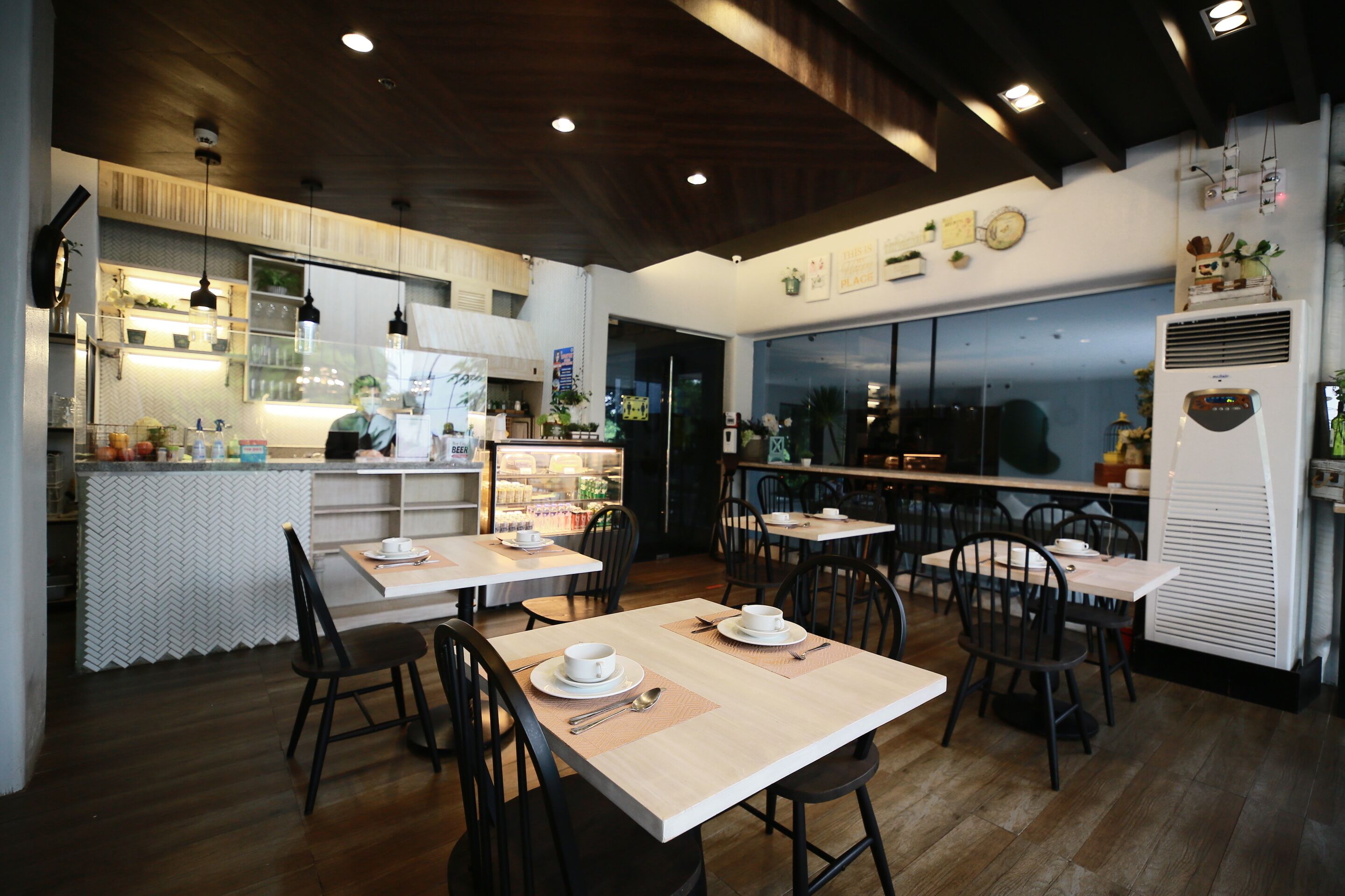 Um restaurante com mesas e cadeiras de madeira, uma área de cozinha e uma janela grande.