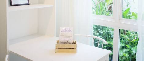Una mesa blanca con un menú enmarcado y una pequeña caja de madera, una silla blanca y un ventanal con vistas a la vegetación.