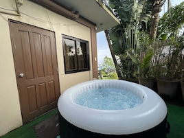 An outdoor hot tub with a wooden door and window, surrounded by potted plants and a bamboo fence.
