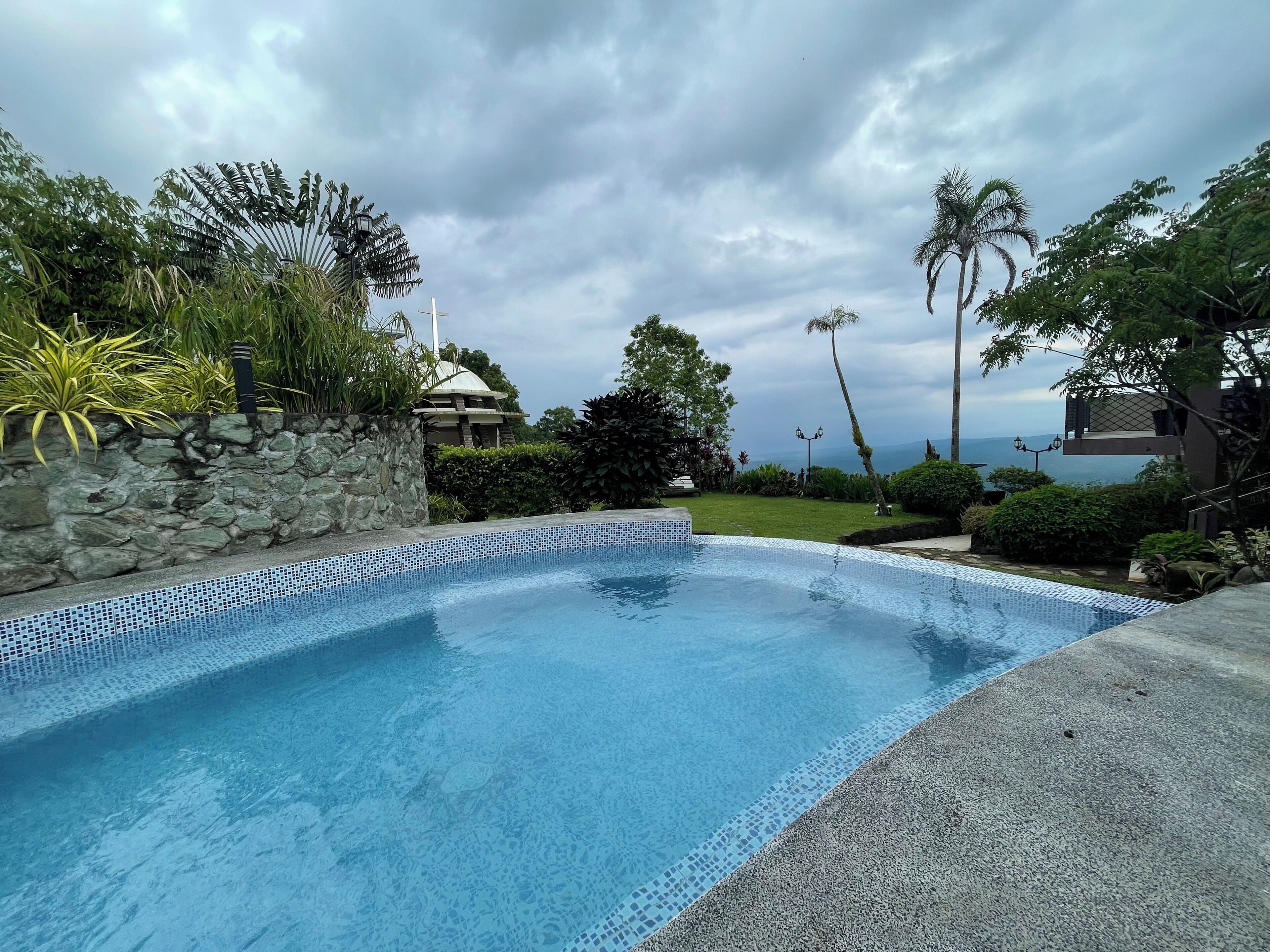 A swimming pool with a stone wall and tiled edge, surrounded by lush greenery and a view of the ocean.