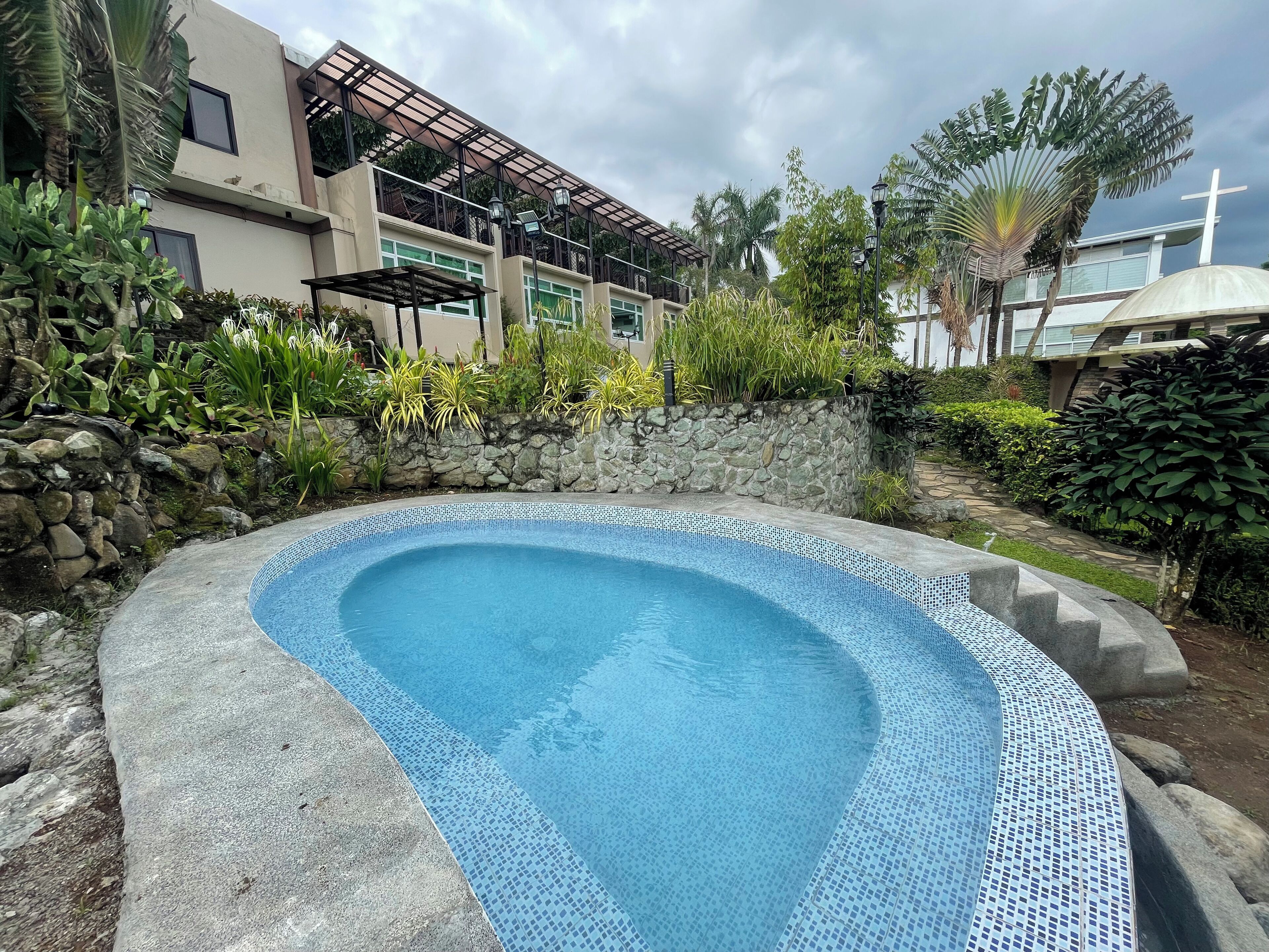 A swimming pool with a stone border, surrounded by lush greenery and a modern building with a cross.