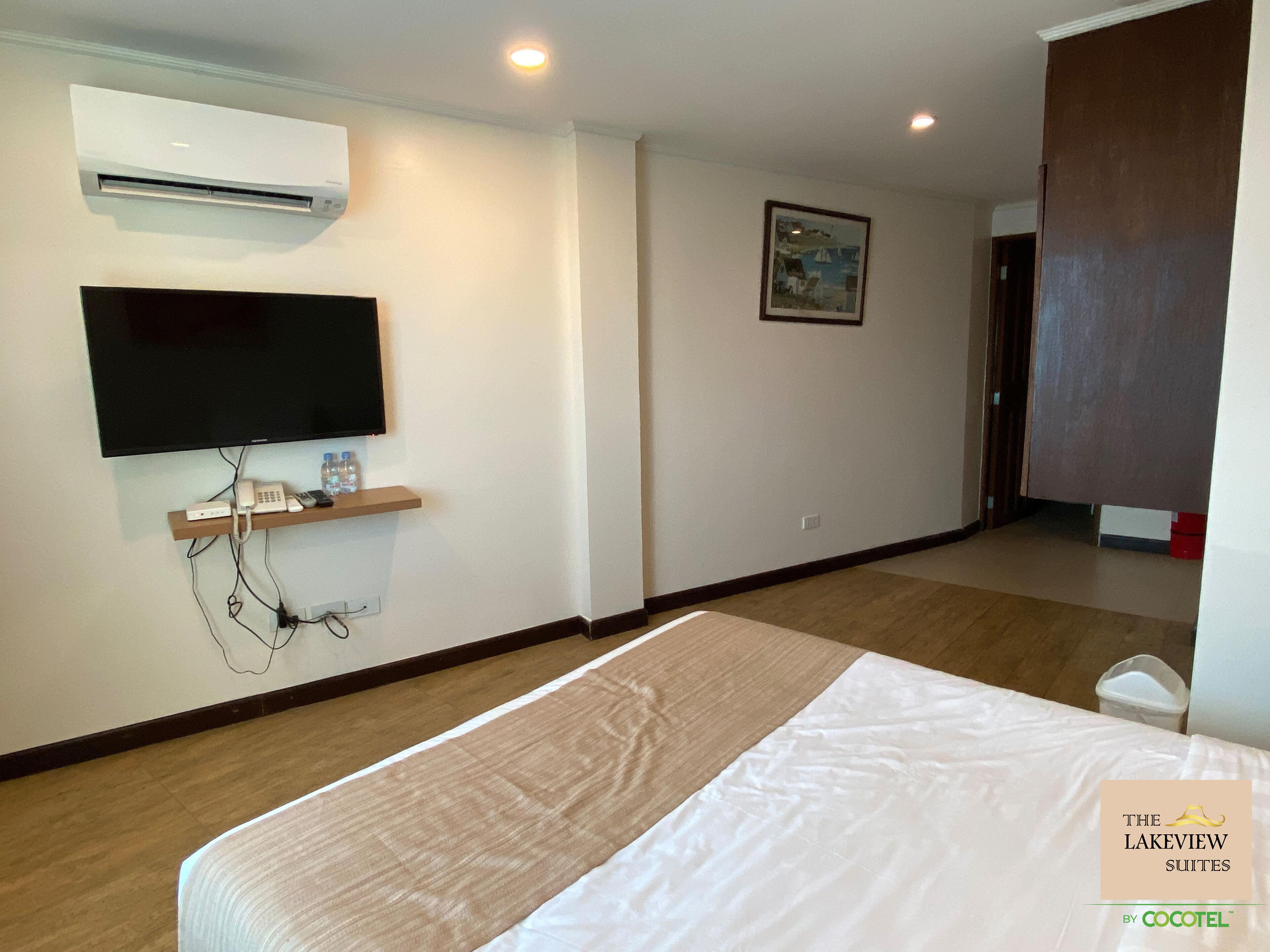 A hotel room with a bed, a mounted TV, a shelf with a phone and water bottles, and a painting on the wall.