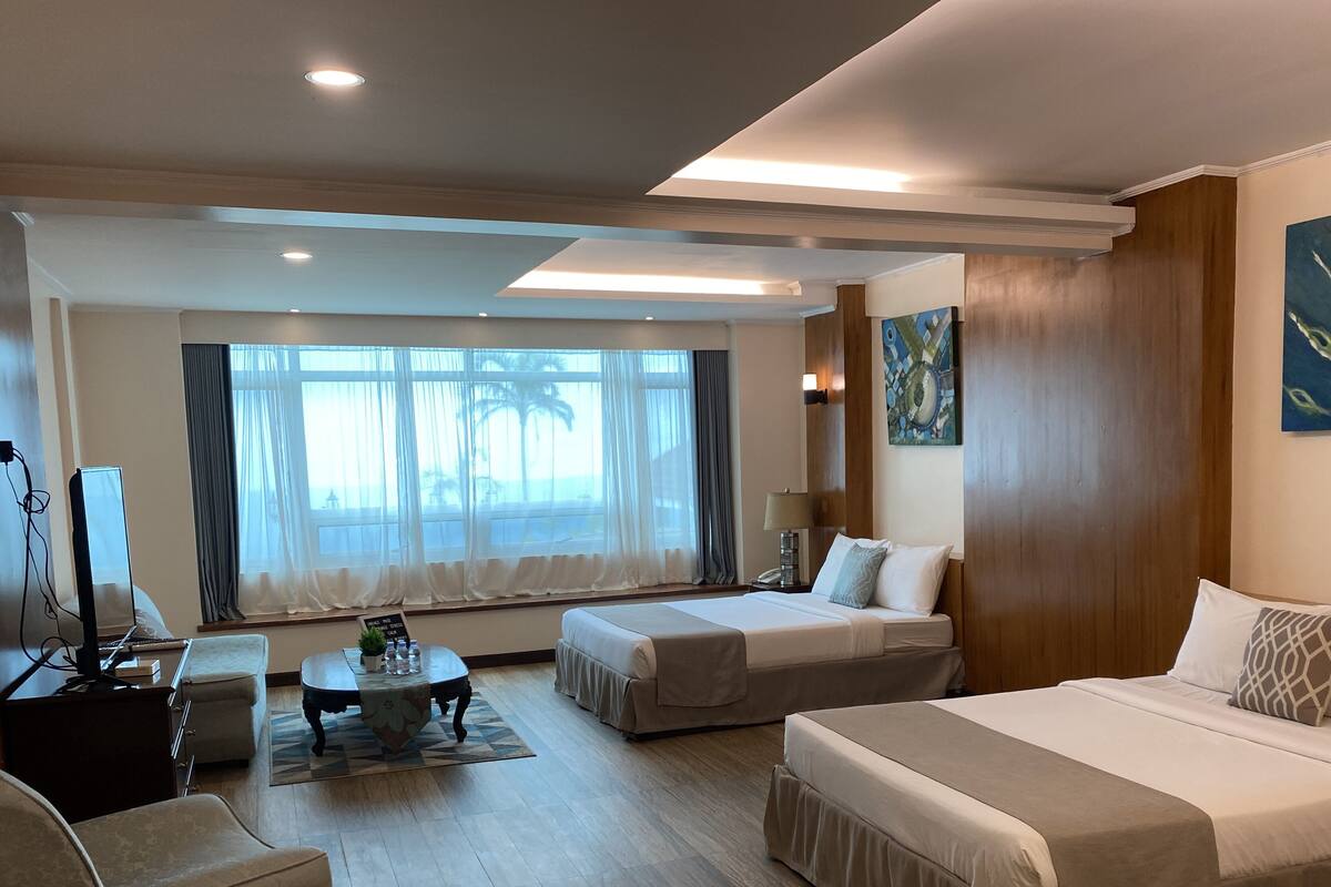 Habitación de hotel con dos camas, un escritorio, una silla, una mesita de centro y un amplio ventanal con cortinas.