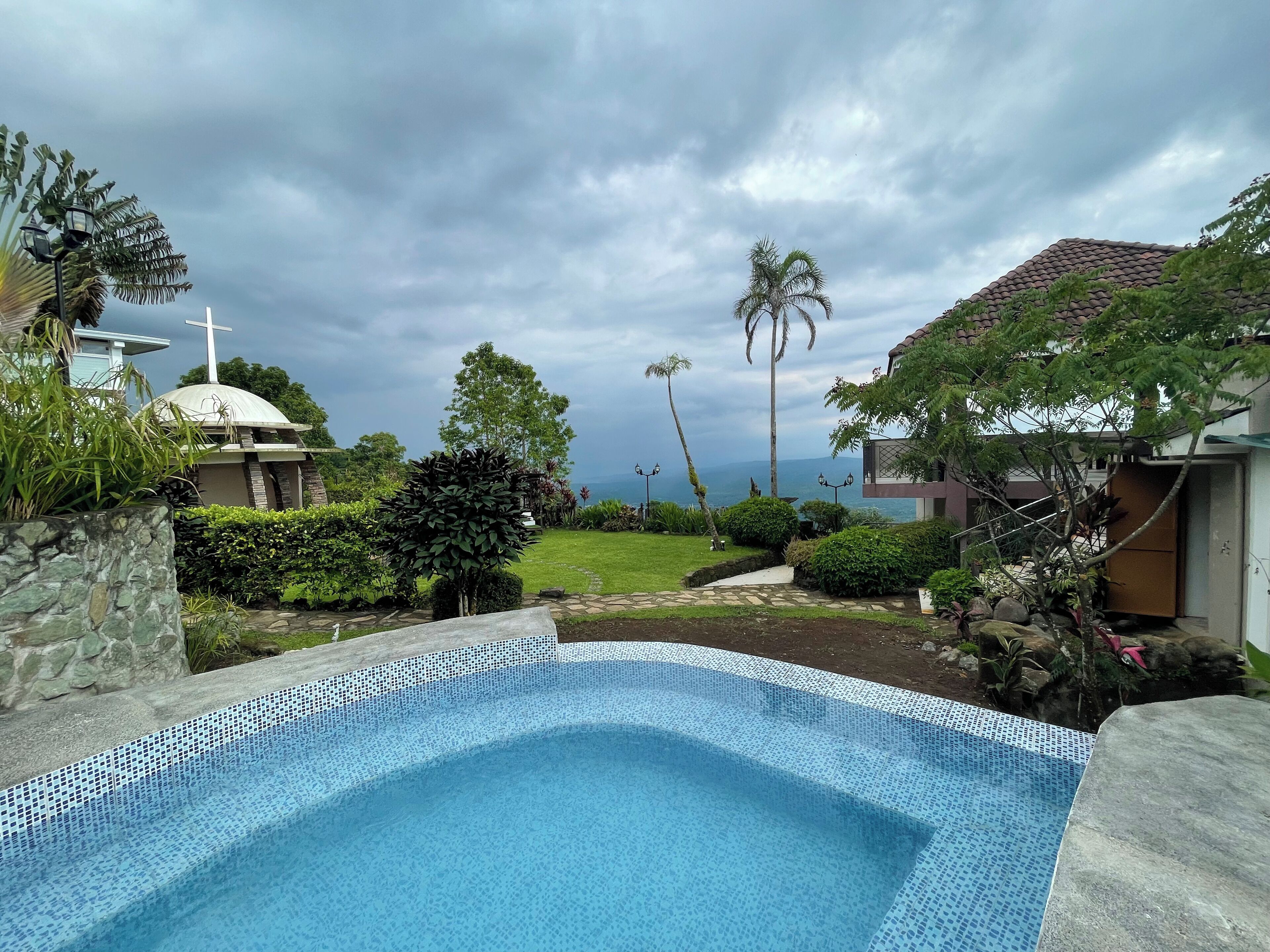 Una piscina con borde de azulejos, rodeada de vegetación exuberante y vistas a una iglesia con cruz.