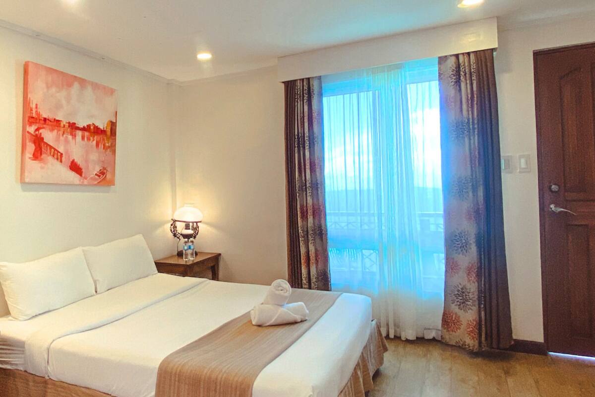 Habitación de hotel con una cama grande, una mesita de noche, una lámpara, un cuadro en la pared y vistas al exterior.