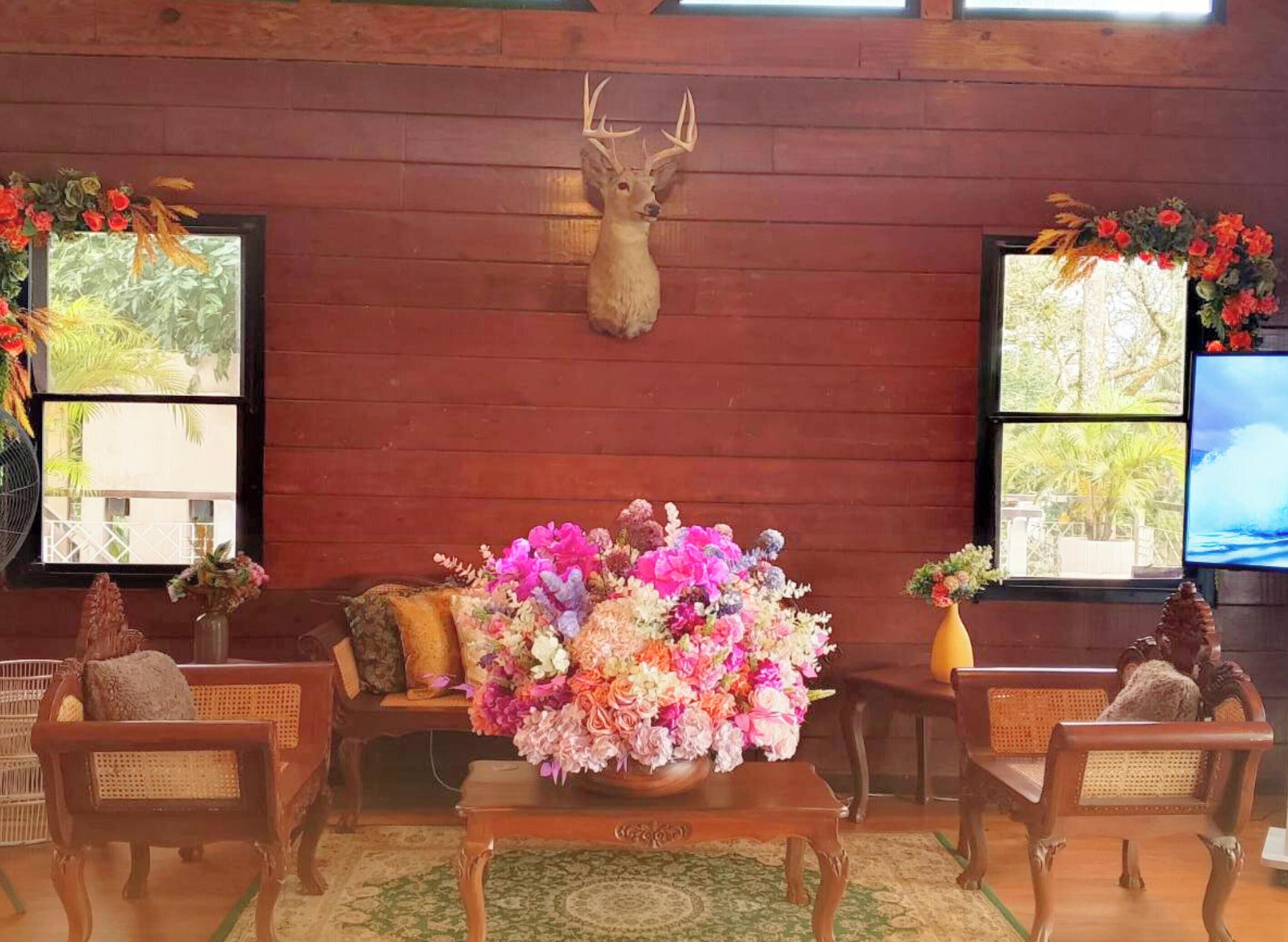 Interior de madera con un ciervo montado en la pared, un centro de mesa floral sobre una mesa, muebles de mimbre y ventanas con vistas.