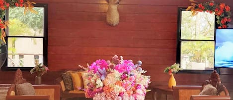 Interior de madera con un ciervo montado en la pared, un centro de mesa floral sobre una mesa, muebles de mimbre y ventanas con vistas.