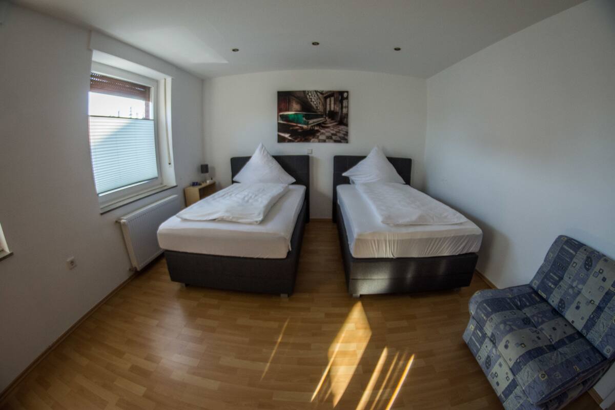 Un dormitorio con dos camas, una ventana con persianas, un radiador, un cuadro en la pared y una silla estampada.