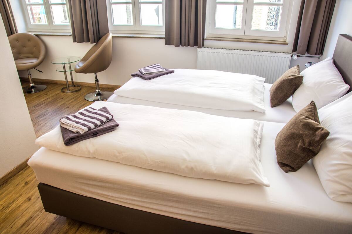 Habitación de hotel con dos camas, una zona de estar con una silla y una mesita, y una ventana con cortinas.