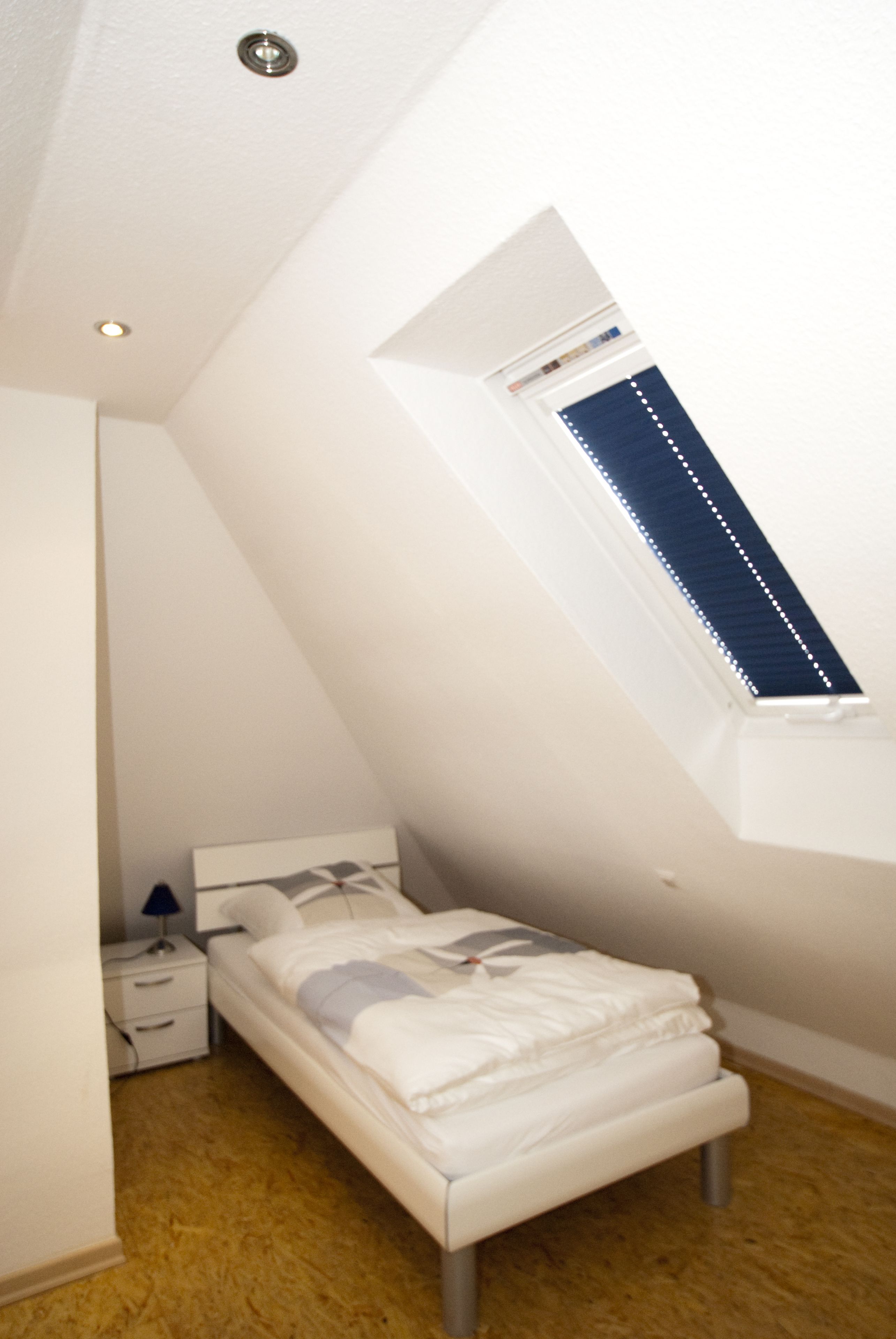 A small, white, sloped-roof bedroom with a single bed, a nightstand, and a window with blinds.