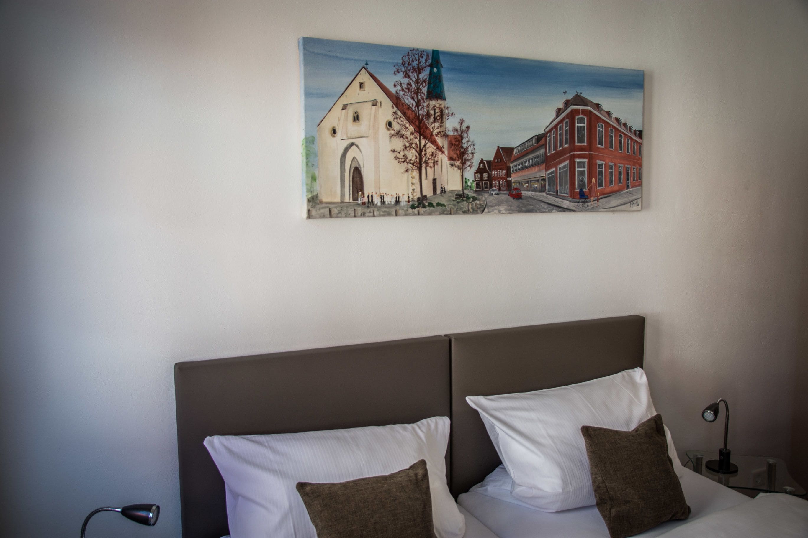 Ein Schlafzimmer mit Bett, Kissen, einem Nachttisch, einer Lampe und einem Bild von einem Gebäude an der Wand.