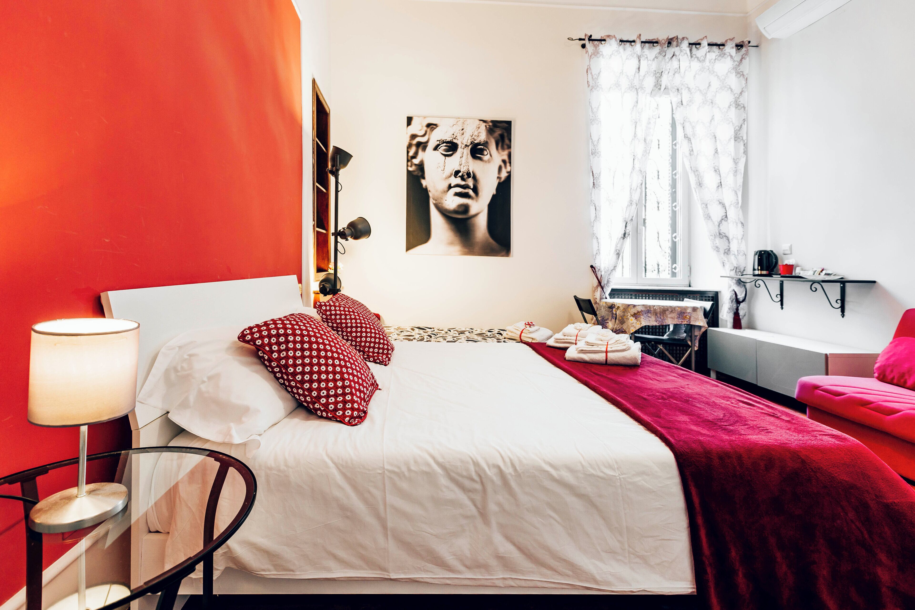 A bedroom with a bed, pillows, a bedside table, a lamp, a window with curtains, and a wall-mounted artwork.