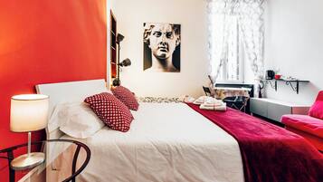 A bedroom with a bed, pillows, a bedside table, a lamp, a window with curtains, and a wall-mounted artwork.