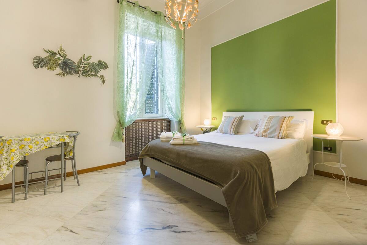 Een slaapkamer met een bed, een tafel met stoelen, een raam met gordijnen, en een groene accentmuur.