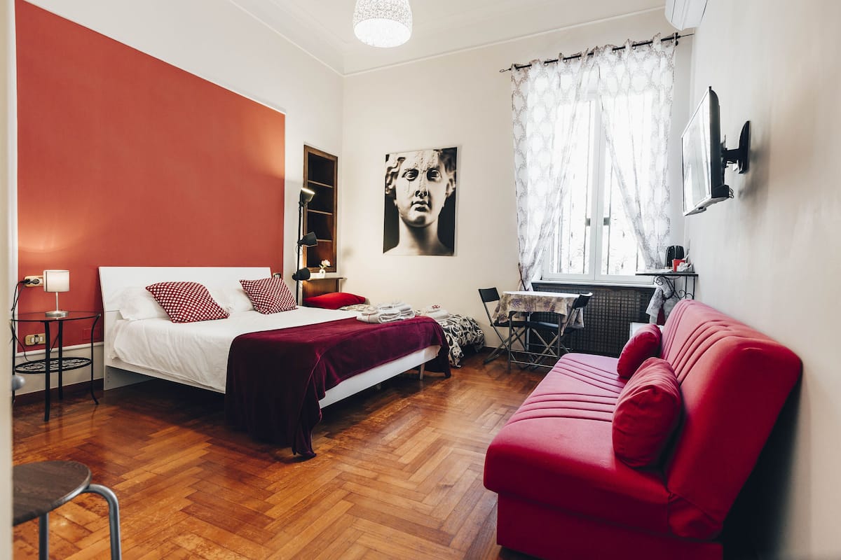 Een slaapkamer met een bed, een rode bank, een bureau met stoel, en een raam met gordijnen.