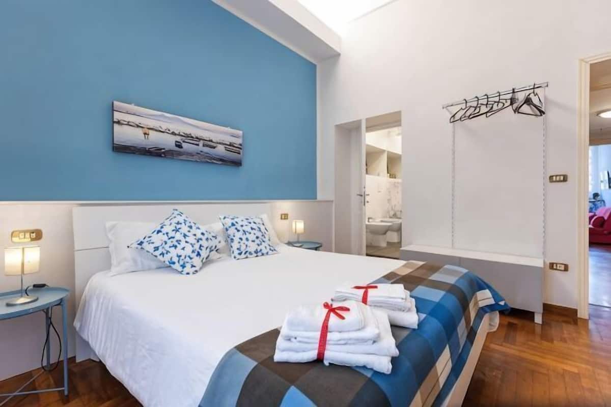 Een slaapkamer met een bed, kussens, een nachtkastje, een aan de muur hangend schilderij en een kledingrek.