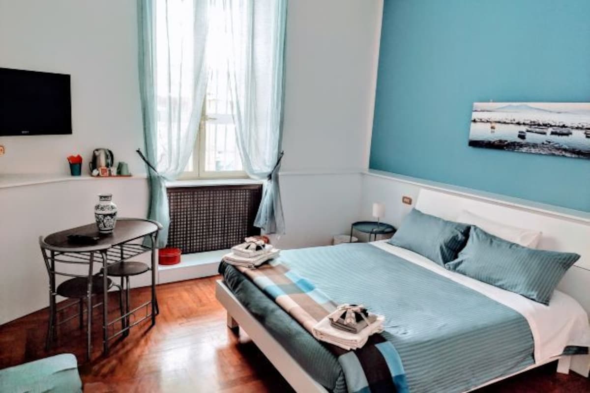 A bedroom with a bed, a small table, a chair, a window with curtains, and a wall-mounted TV.