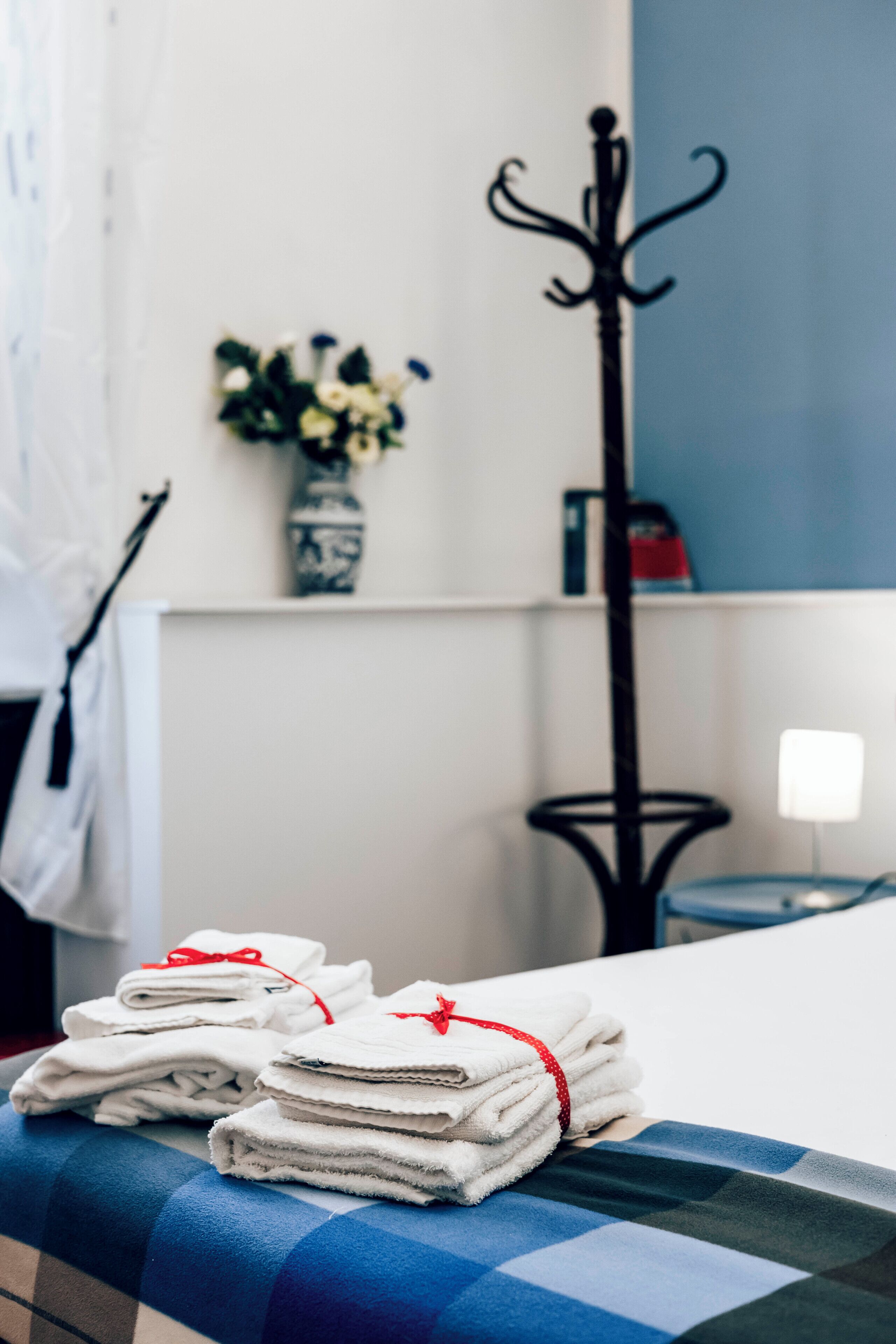 A neatly folded stack of white towels tied with a red ribbon on a checkered bedspread, a black metal coat rack, and a vase with flowers on a white wall.