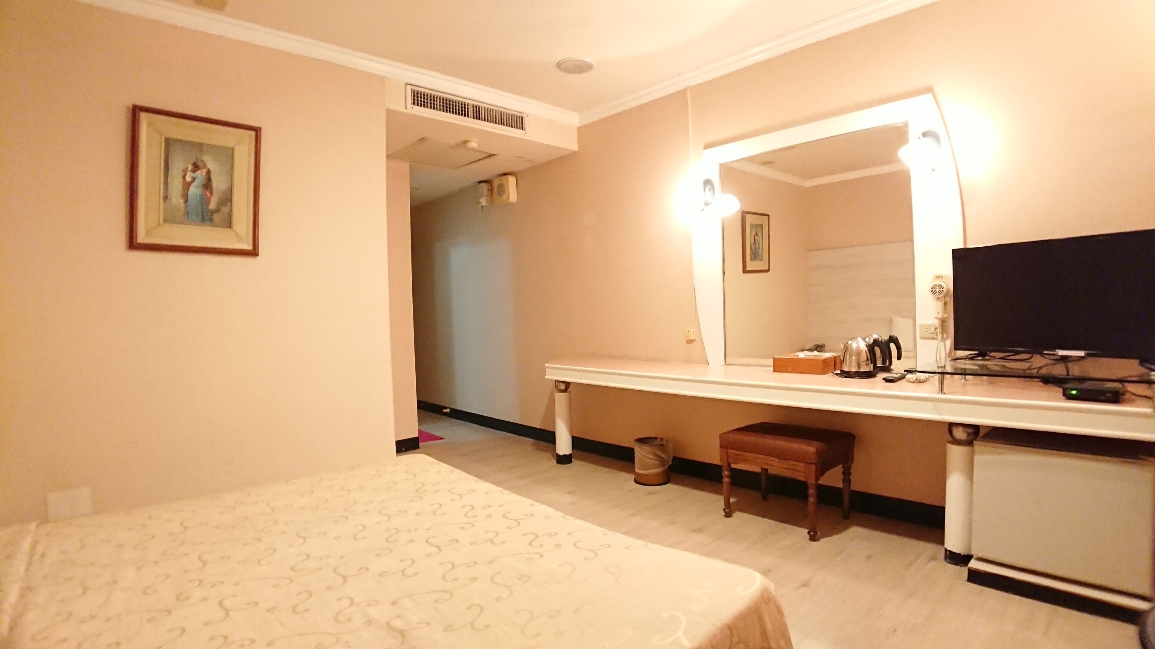 A hotel room with a bed, a desk with a mirror, a television, and a small stool.
