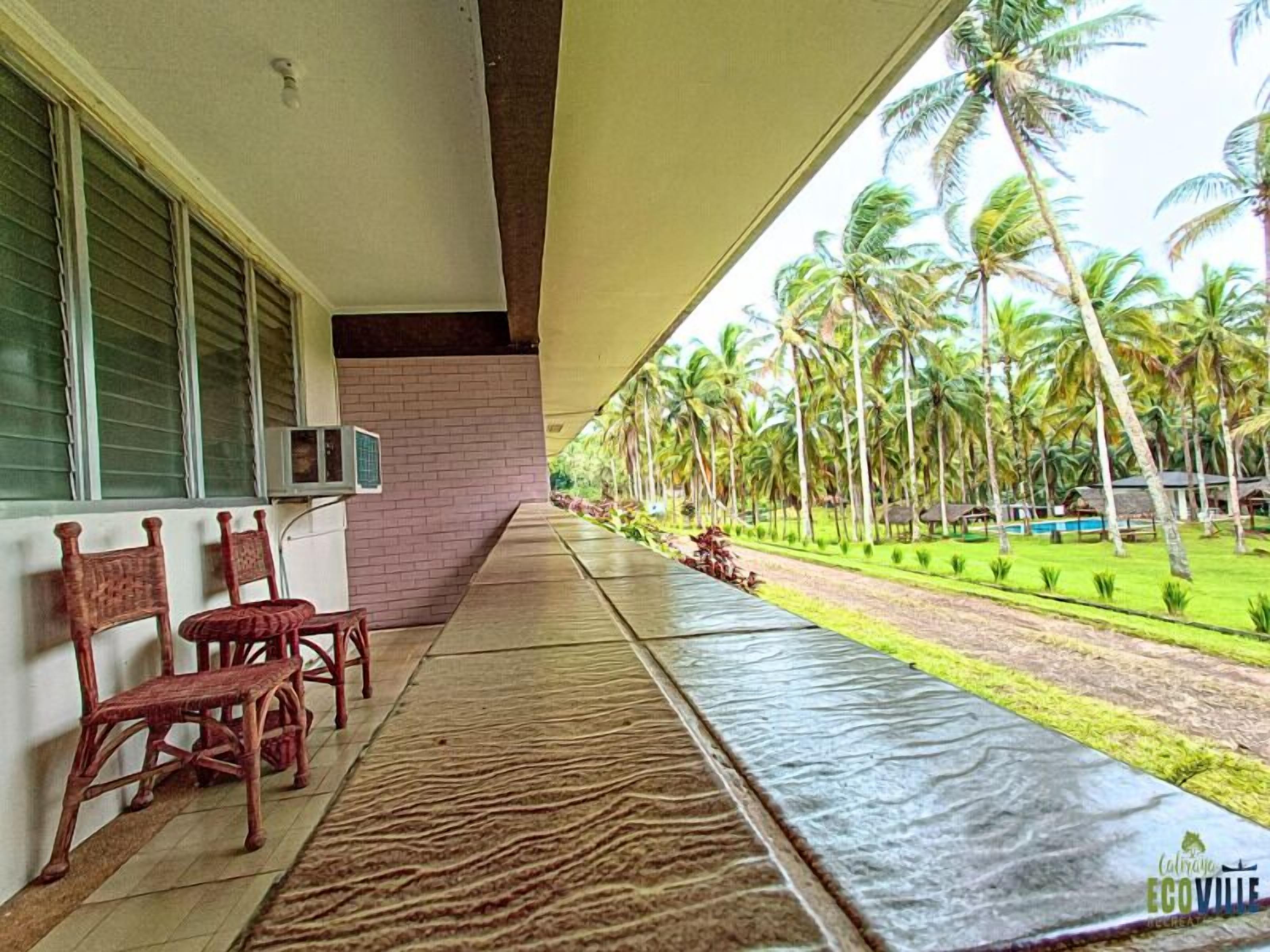 A covered outdoor area with a view of palm trees and a pool. There are wooden chairs and a brick wall.