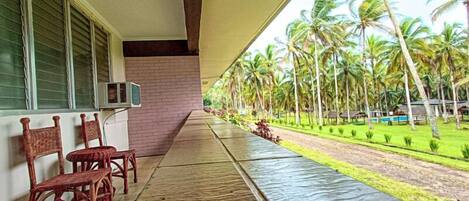 Un espacio al aire libre techado con vista a palmeras y una piscina. Se observan sillas de madera y una pared de ladrillo.