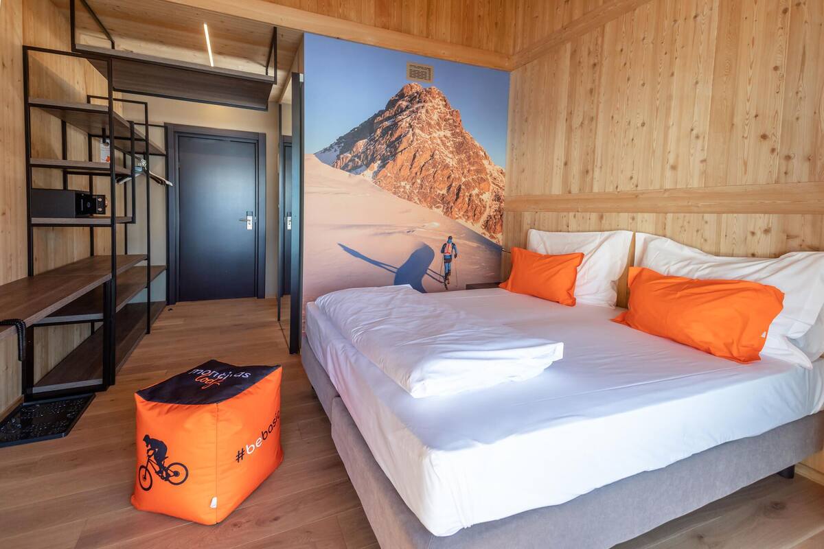 A room with a bed, a wooden desk, a chair, and a wall with a mountain painting.