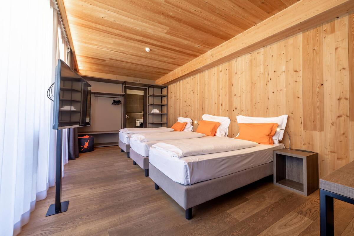 A hotel room with a wooden ceiling, a bed with white and orange bedding, a small table, and a TV.