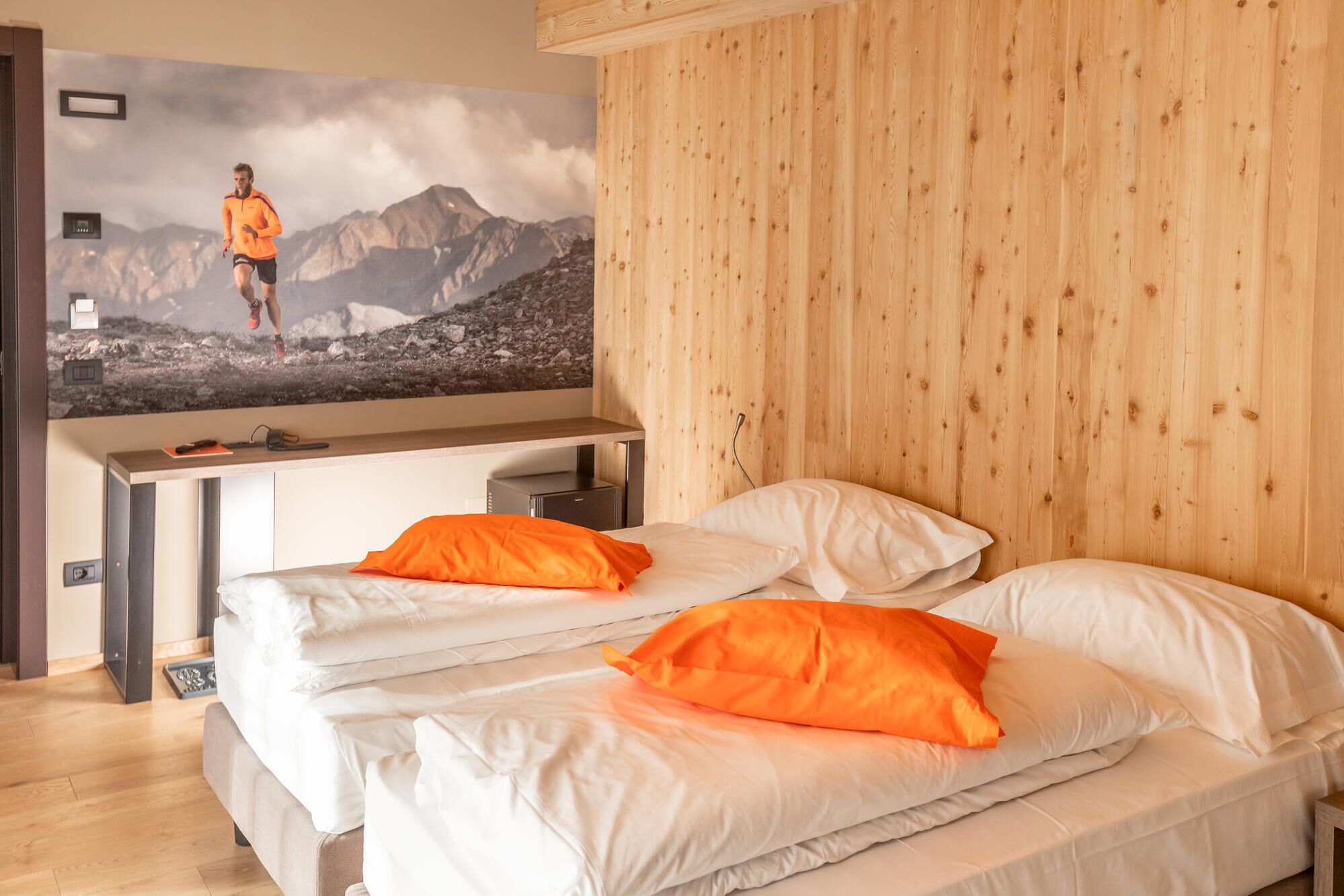Habitació d'hotel amb llit, dos travessers, escriptori, cadira i televisió penjada a la paret que mostra un corredor.
