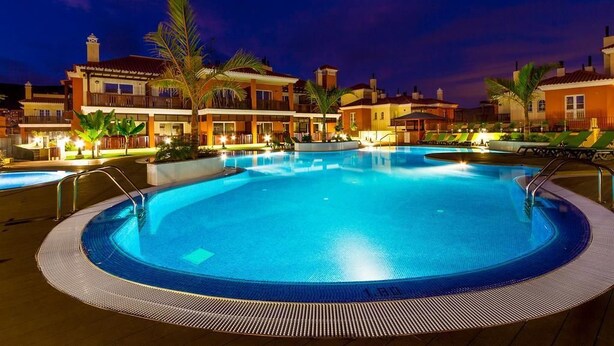 Una piscina rodeada de edificios con balcones, palmeras y un cielo nocturno.